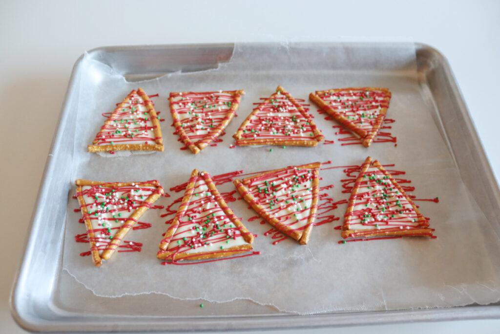 Eight triangle-shaped Christmas cookies decorated with red icing and green sprinkles are arranged on a parchment-lined baking sheet.