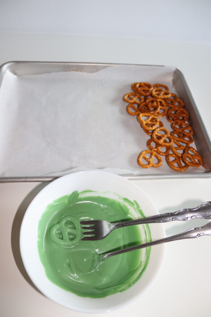 A baking sheet with parchment paper holds pretzels in the background; a bowl with green melted coating and forks is in the foreground.