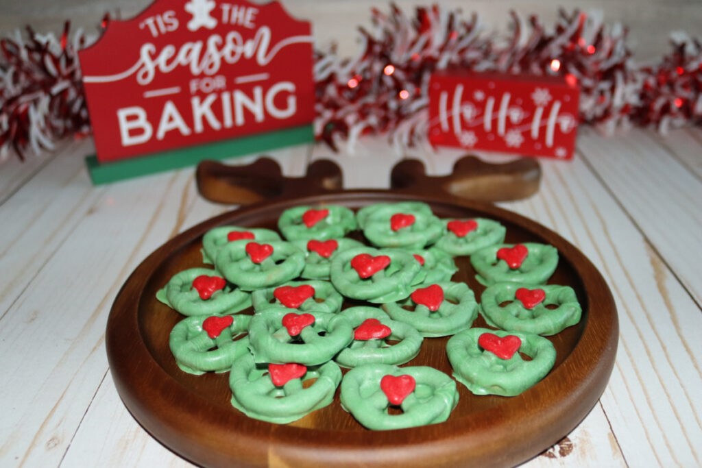 A wooden plate with green Christmas wreath cookies topped with red decorations, set on a white surface with festive signs and garland in the background.