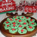 A wooden plate with green Christmas wreath cookies topped with red decorations, set on a white surface with festive signs and garland in the background.
