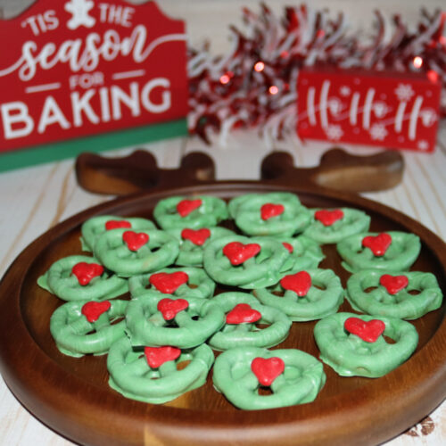A wooden plate with green Christmas wreath cookies topped with red decorations, set on a white surface with festive signs and garland in the background.