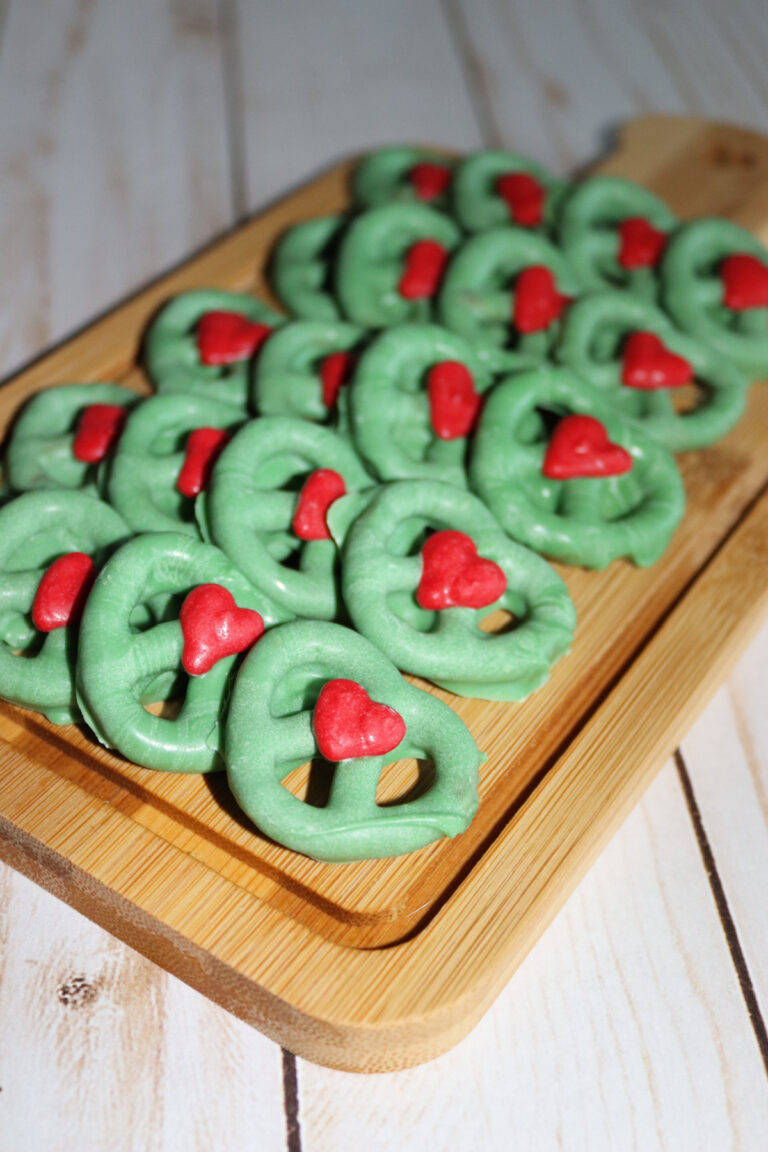 Green pretzels with red heart decorations are arranged on a wooden cutting board atop a light-colored wood surface.