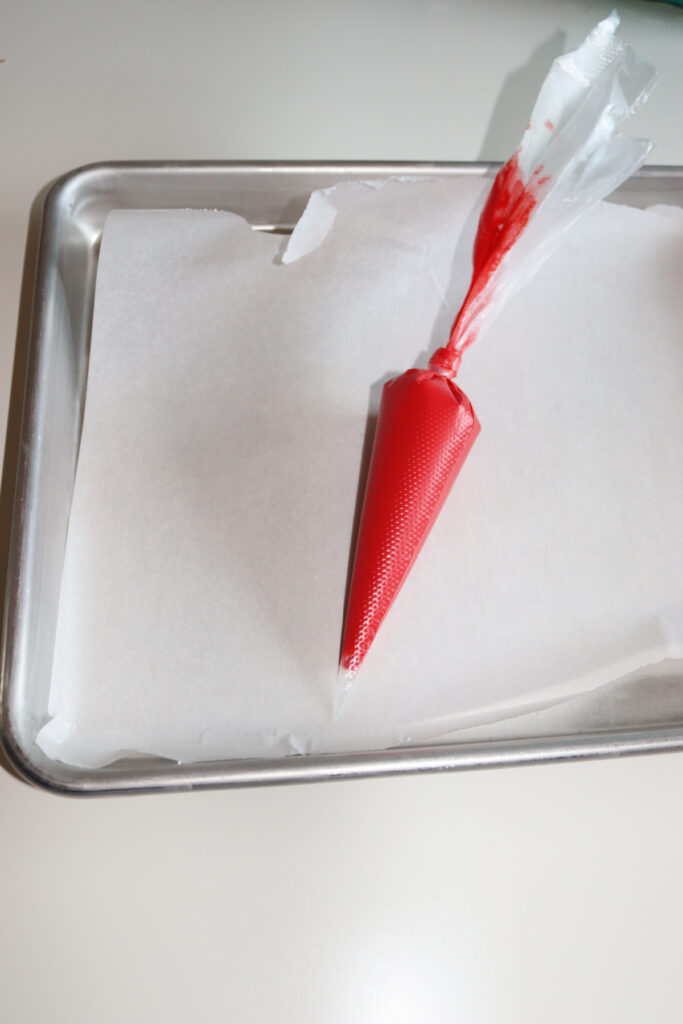 A piping bag filled with red icing rests on a baking tray lined with parchment paper.
