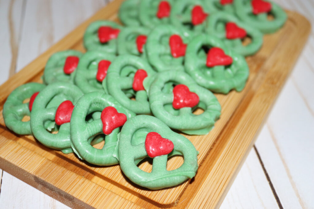 A wooden tray with green pretzels topped with small red heart-shaped candies, arranged in rows on a light-colored surface.