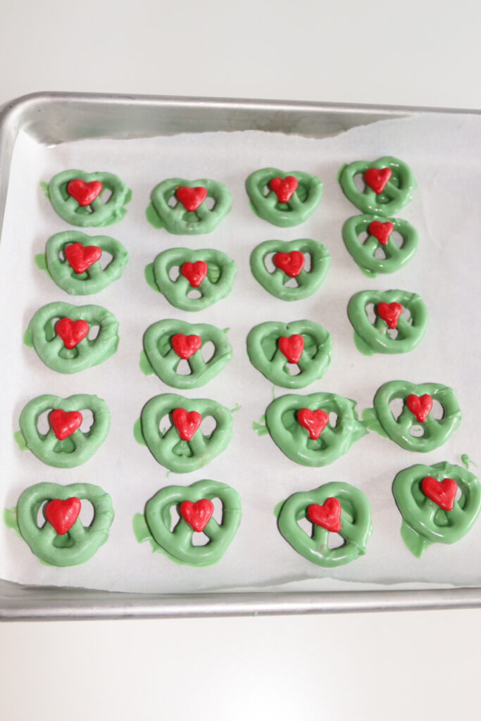 A tray of green candy-coated pretzels, each topped with a red heart-shaped candy, arranged on parchment paper.