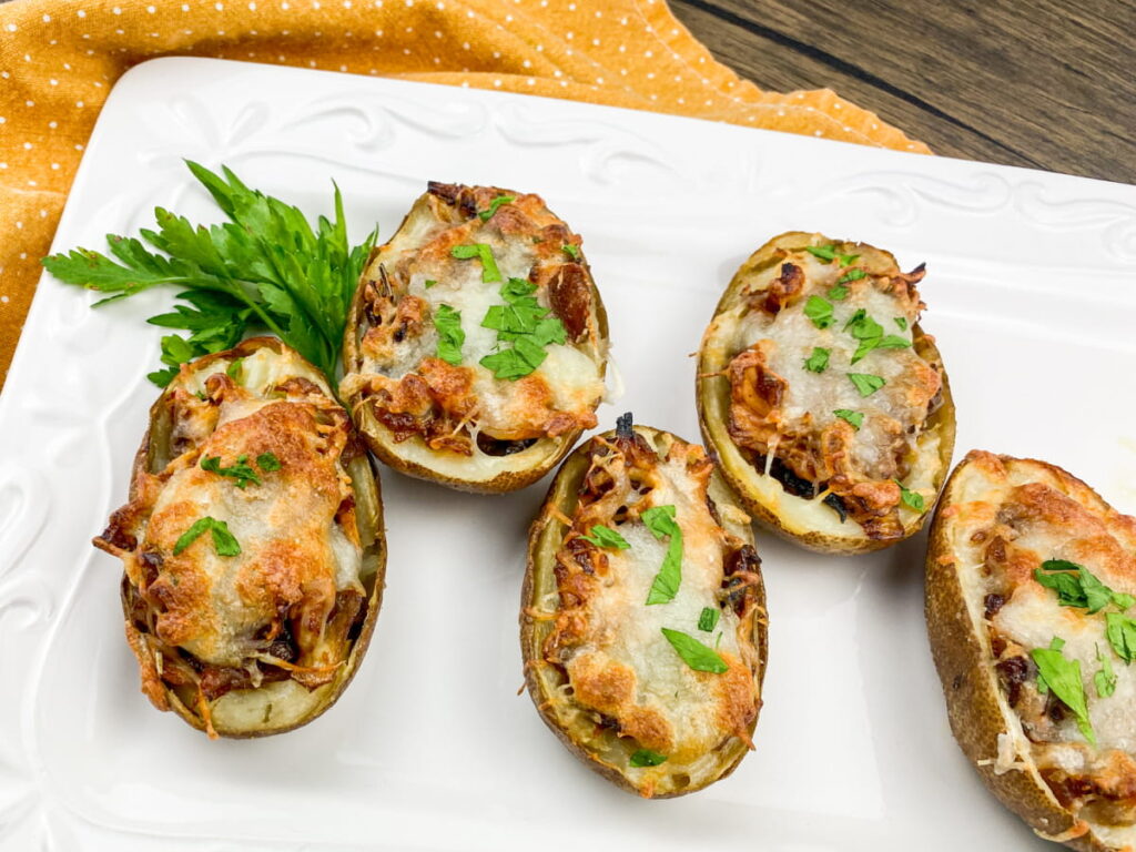 Five baked potato halves topped with melted cheese and herbs are arranged on a white plate, garnished with fresh parsley.