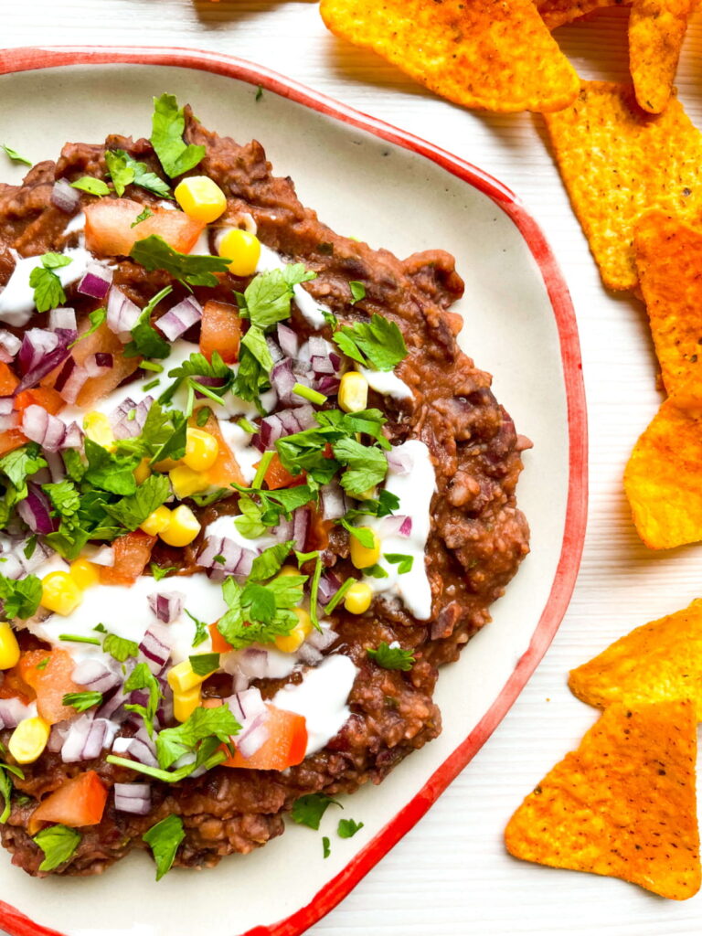 A plate of refried beans topped with chopped tomato, corn, red onion, cilantro, and sour cream, served with tortilla chips on the side.