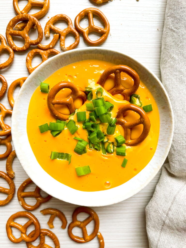 A bowl of cheese dip topped with pretzels and chopped green onions, surrounded by scattered pretzels on a white surface.