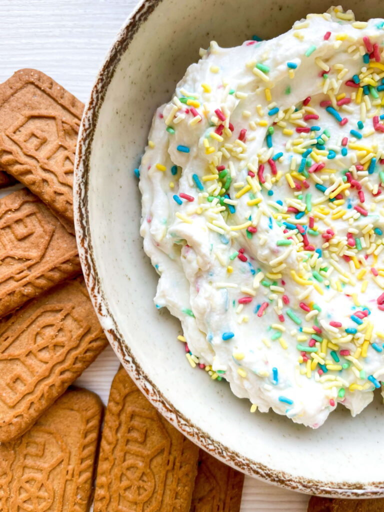 A bowl of creamy dip topped with colorful sprinkles is next to several rectangular biscuits on a white surface.