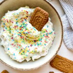 A bowl of whipped dip topped with colorful sprinkles and a rectangular cookie, with more similar cookies placed nearby on a white surface.