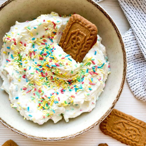 A bowl of whipped dip topped with colorful sprinkles and a rectangular cookie, with more similar cookies placed nearby on a white surface.