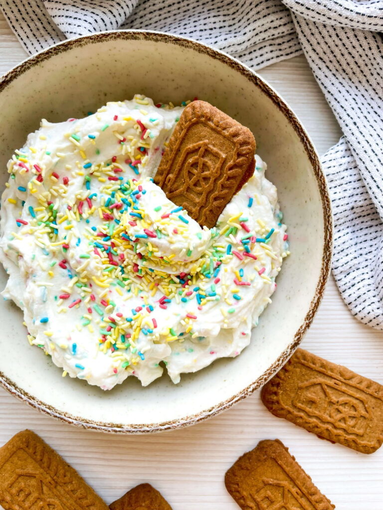 A bowl of whipped dip topped with colorful sprinkles and a rectangular cookie, with more similar cookies placed nearby on a white surface.