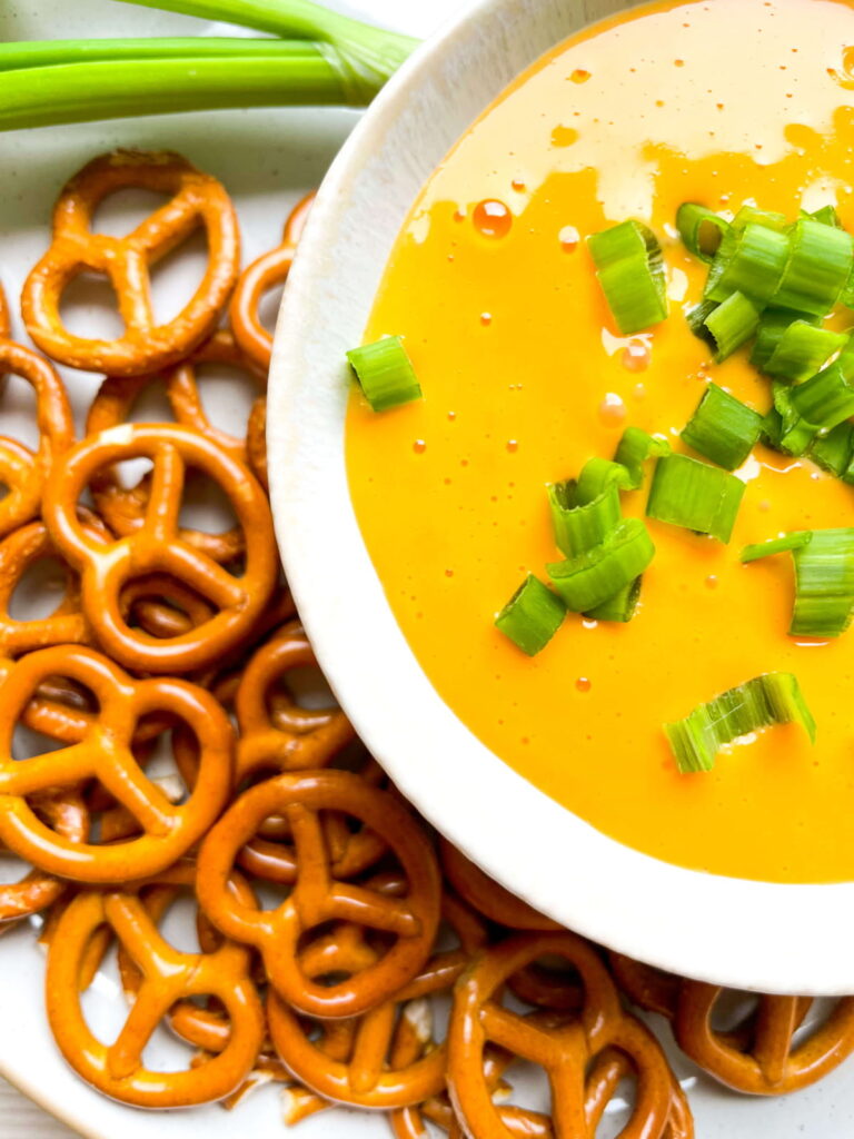 A plate of pretzels next to a bowl of yellow cheese dip topped with chopped green onions.