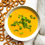 A bowl of yellow cheese dip topped with chopped green onions, surrounded by pretzel twists on a white surface with a beige cloth nearby.