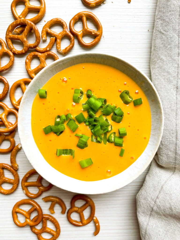 A bowl of yellow cheese dip topped with chopped green onions, surrounded by pretzel twists on a white surface with a beige cloth nearby.