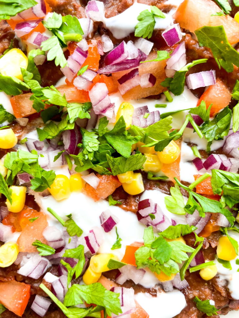 Close-up of a layered dip topped with chopped red onions, tomatoes, corn, cilantro, and a drizzle of white sauce.