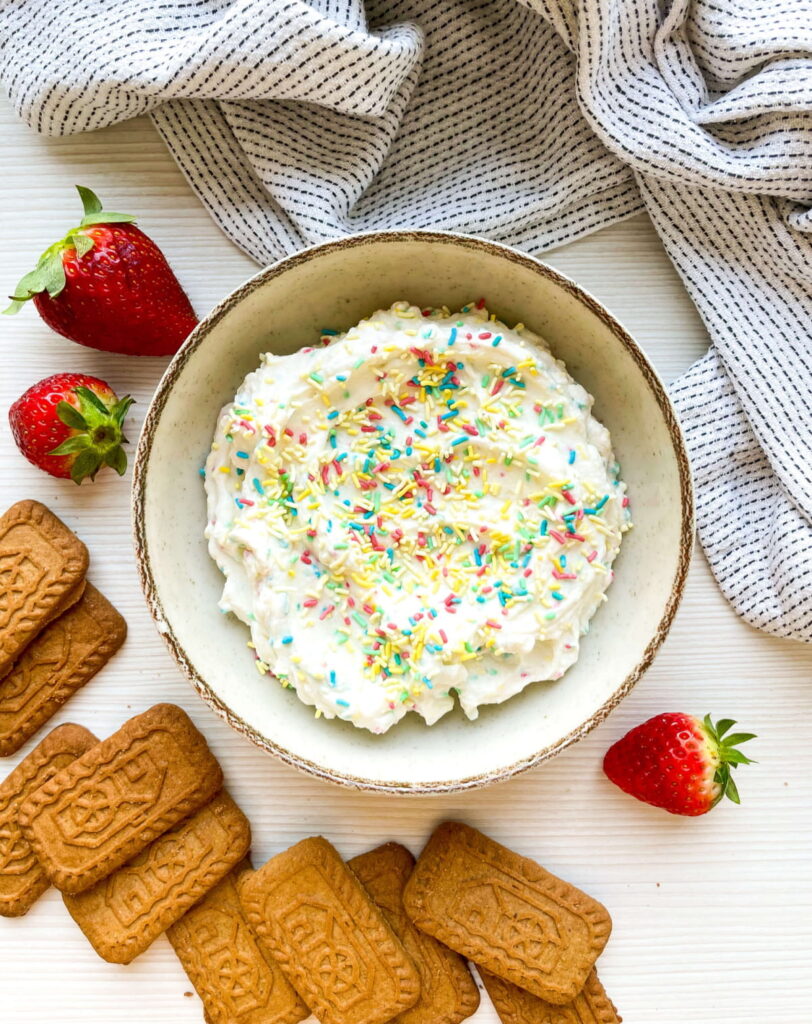 A bowl of white cream dip topped with colorful sprinkles, surrounded by strawberries and rectangular brown cookies, placed on a white surface with a striped towel nearby.
