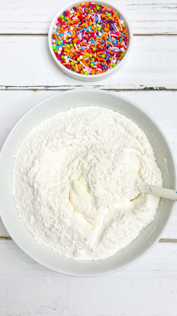A bowl of flour mixture with a spatula next to a small bowl of colorful sprinkles on a white wooden surface.