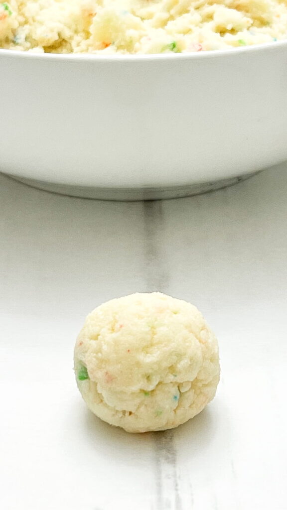 A ball of cookie dough with colored sprinkles sits on a white surface, with a bowl of more dough in the background.