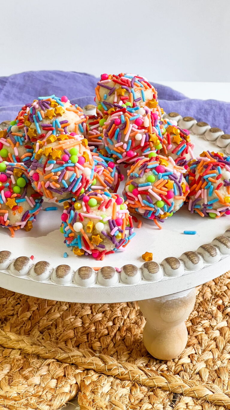 A plate of round cake balls covered in colorful sprinkles is displayed on a white stand with a woven mat underneath.