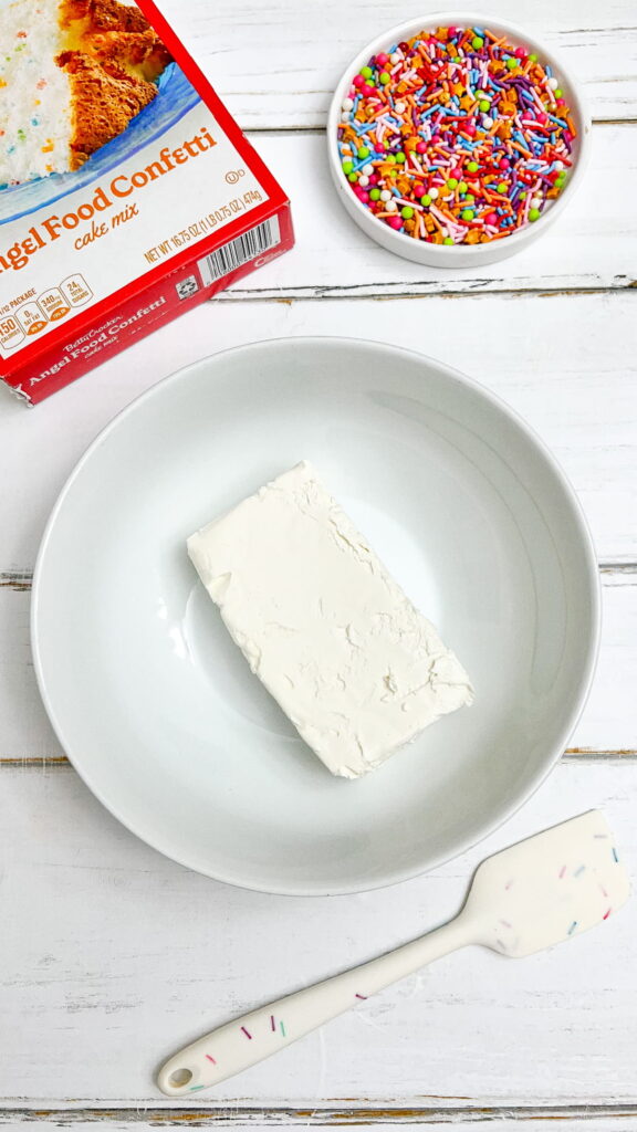 A block of cream cheese in a white bowl, a box of angel food confetti cake mix, a bowl of rainbow sprinkles, and a spatula on a white wooden surface.