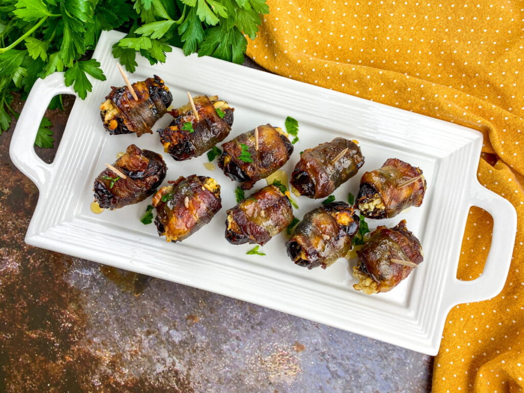 A rectangular white platter holds twelve bacon-wrapped dates stuffed with cheese and herbs, placed beside fresh parsley and a yellow dotted cloth.