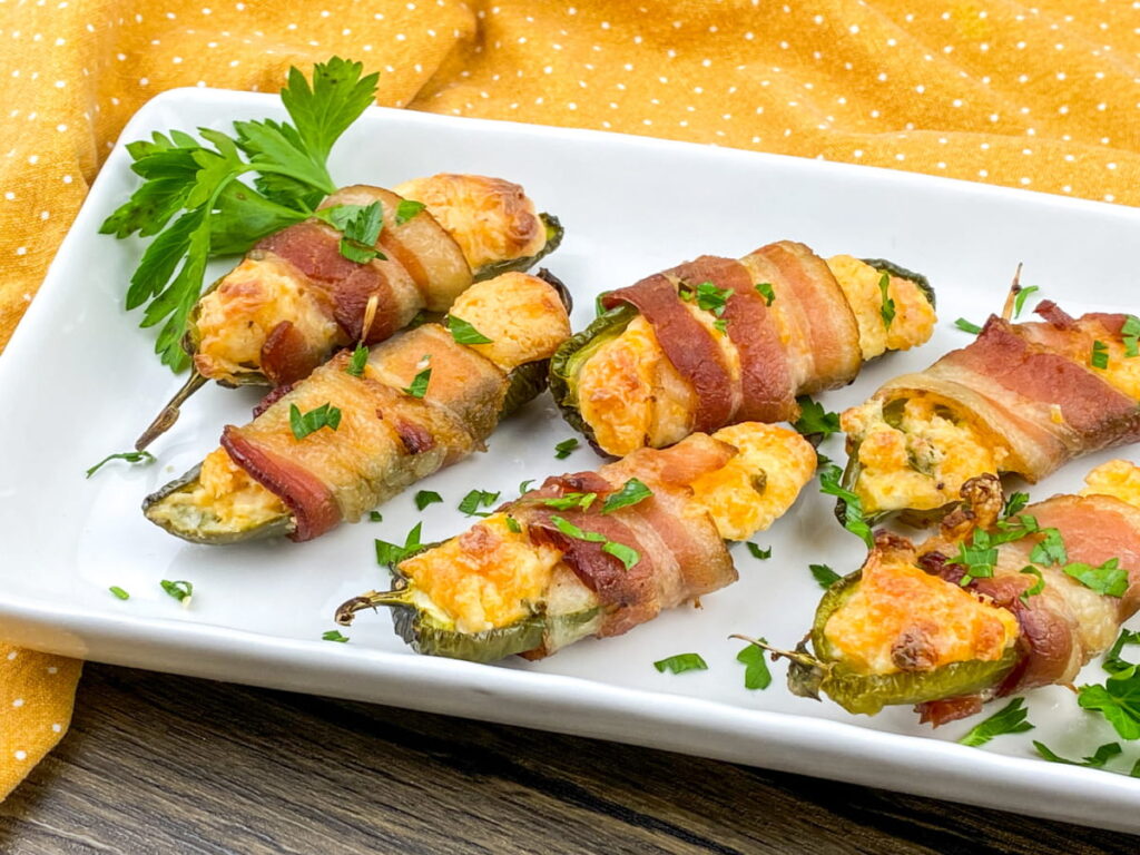 Bacon-wrapped jalapeño poppers stuffed with cheese are arranged on a white rectangular plate, garnished with chopped parsley.