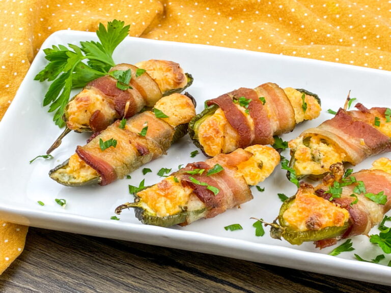 Bacon-wrapped jalapeño poppers stuffed with cheese are arranged on a white rectangular plate, garnished with chopped parsley.