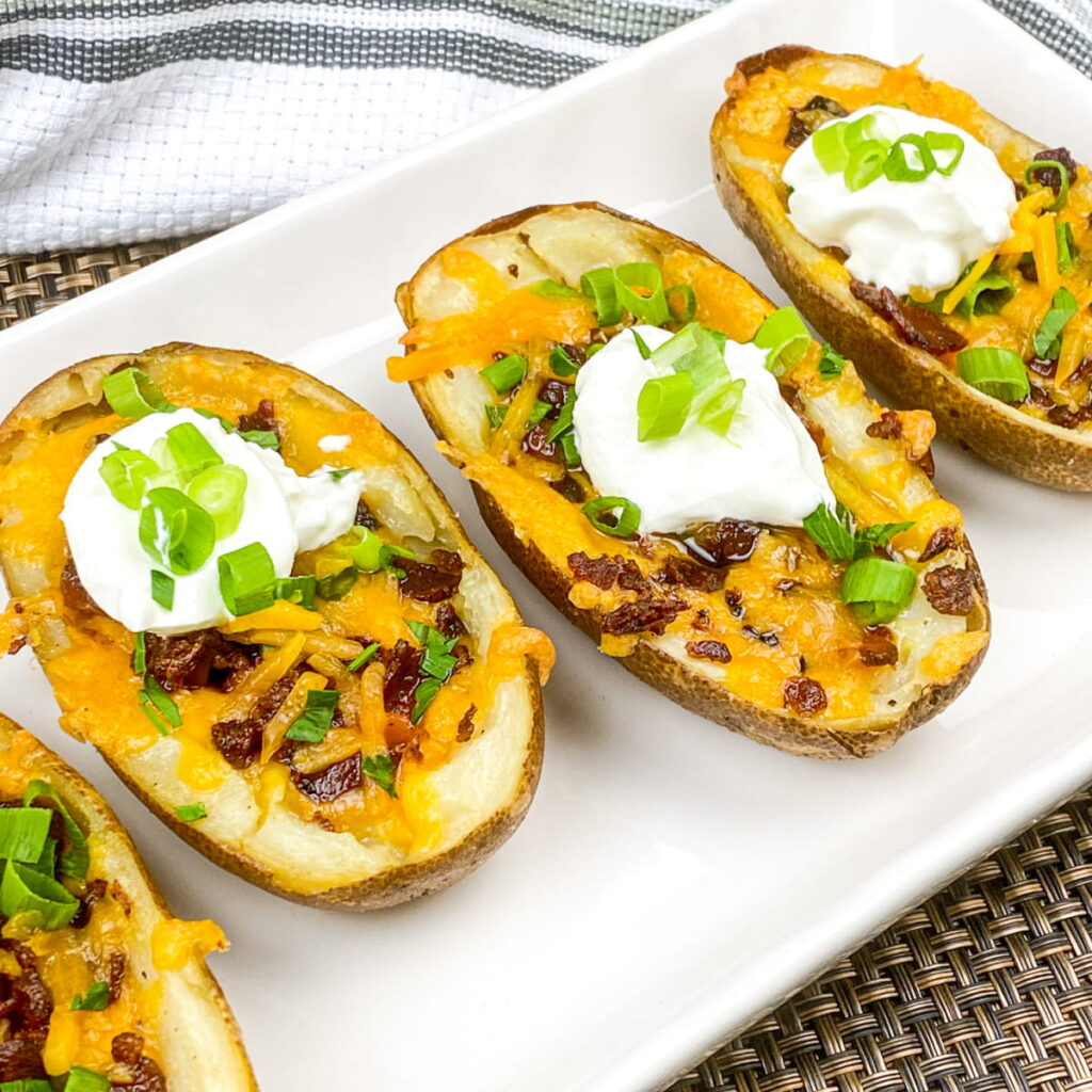 Four potato skins on a white plate, topped with melted cheese, bacon bits, sour cream, and chopped green onions.