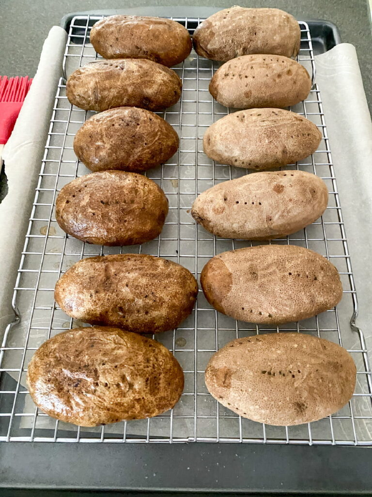 Eleven whole baked potatoes cooling on a wire rack set over a baking sheet.