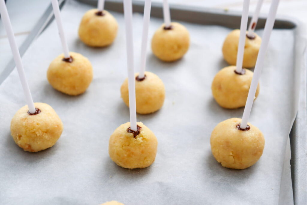 Baking tray with round cake pops on sticks, each placed on parchment paper and secured with a small amount of chocolate at the base of the stick.