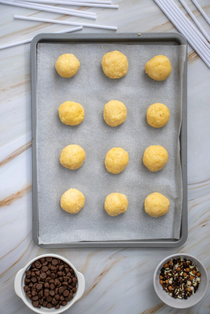 A baking tray lined with parchment paper holds twelve evenly spaced balls of dough; bowls with chocolate chips and chopped nuts are nearby.