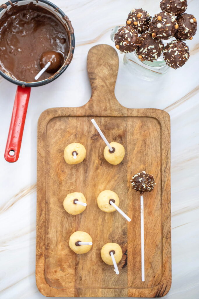 A wooden board holds six uncoated cake pops on sticks; nearby are a jar of finished chocolate-coated cake pops and a saucepan with melted chocolate.