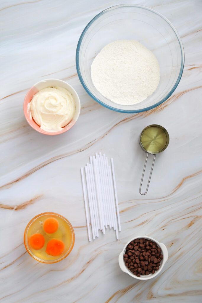 Five baking ingredients—flour, cream cheese, eggs, chocolate chips, a metal measuring cup—and a pile of white sticks arranged on a marble surface.