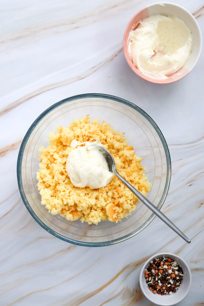 A glass bowl with crumbled cake and a dollop of frosting, a spoon, a small bowl of frosting, and a bowl of colorful sprinkles on a marble surface.
