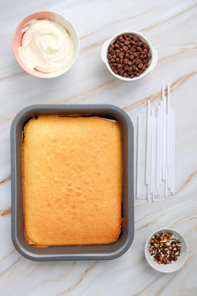 A baked rectangular cake in a pan, surrounded by bowls of white frosting, chocolate chips, sprinkles, and white cake pop sticks on a marble surface.