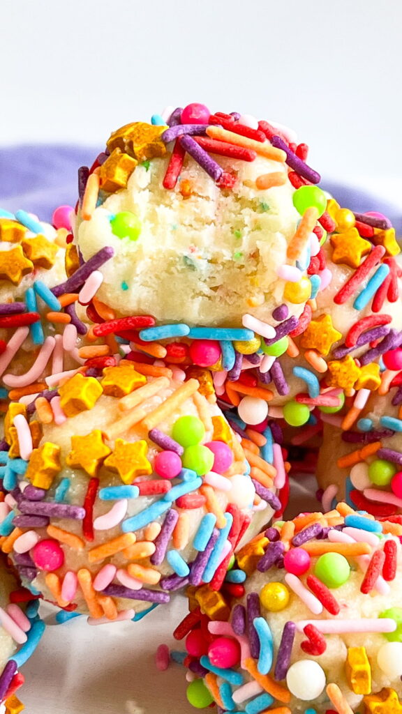 A close-up of colorful sprinkle-covered cookie dough balls, with one showing a bite taken out, revealing the soft interior.