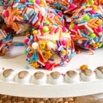 Colorful sprinkle-covered cake balls arranged on a white cake stand with a textured woven mat underneath.