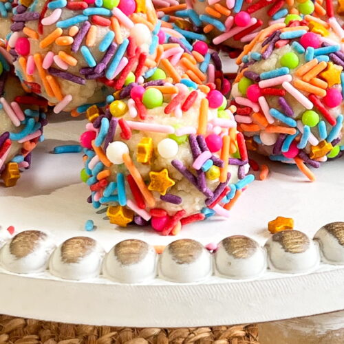 Colorful sprinkle-covered cake balls arranged on a white cake stand with a textured woven mat underneath.