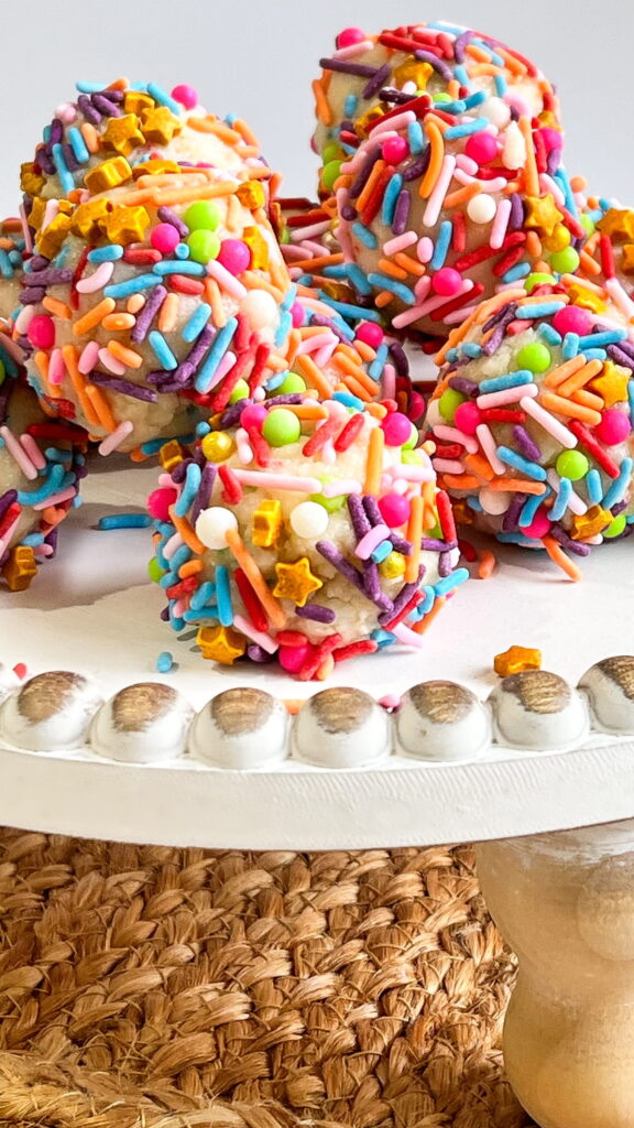 Colorful sprinkle-covered cake balls arranged on a white cake stand with a textured woven mat underneath.