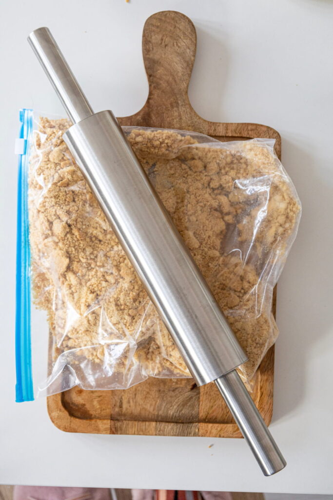 A metal rolling pin rests on a wooden cutting board with a resealable bag of crushed crackers underneath.