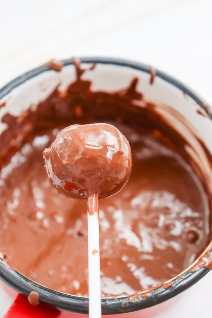 A cake pop on a stick is coated in melted chocolate, held above a bowl filled with more melted chocolate.