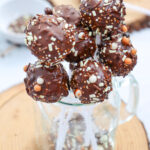 A cluster of chocolate-covered cake pops with sprinkles is arranged in a glass jar, set on a wooden surface.