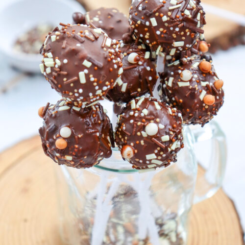 A cluster of chocolate-covered cake pops with sprinkles is arranged in a glass jar, set on a wooden surface.