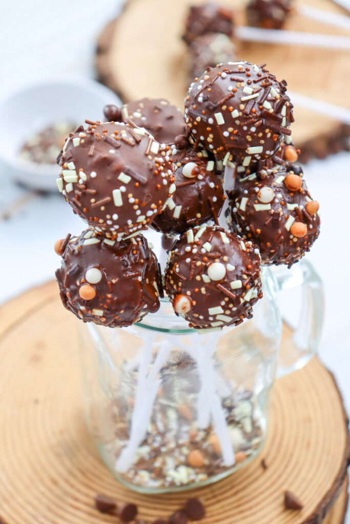 A cluster of chocolate-covered cake pops with sprinkles is arranged in a glass jar, set on a wooden surface.
