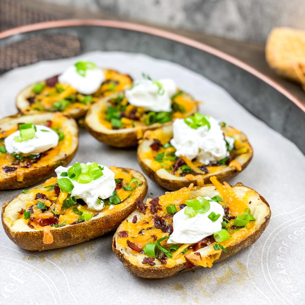 Six potato skins topped with melted cheese, bacon bits, sour cream, and chopped green onions are arranged on parchment paper on a tray.