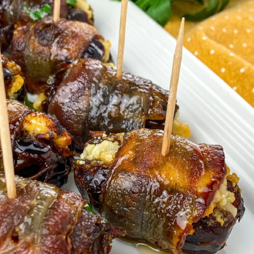 Bacon-wrapped stuffed dates secured with toothpicks are arranged on a white plate, garnished with herbs.