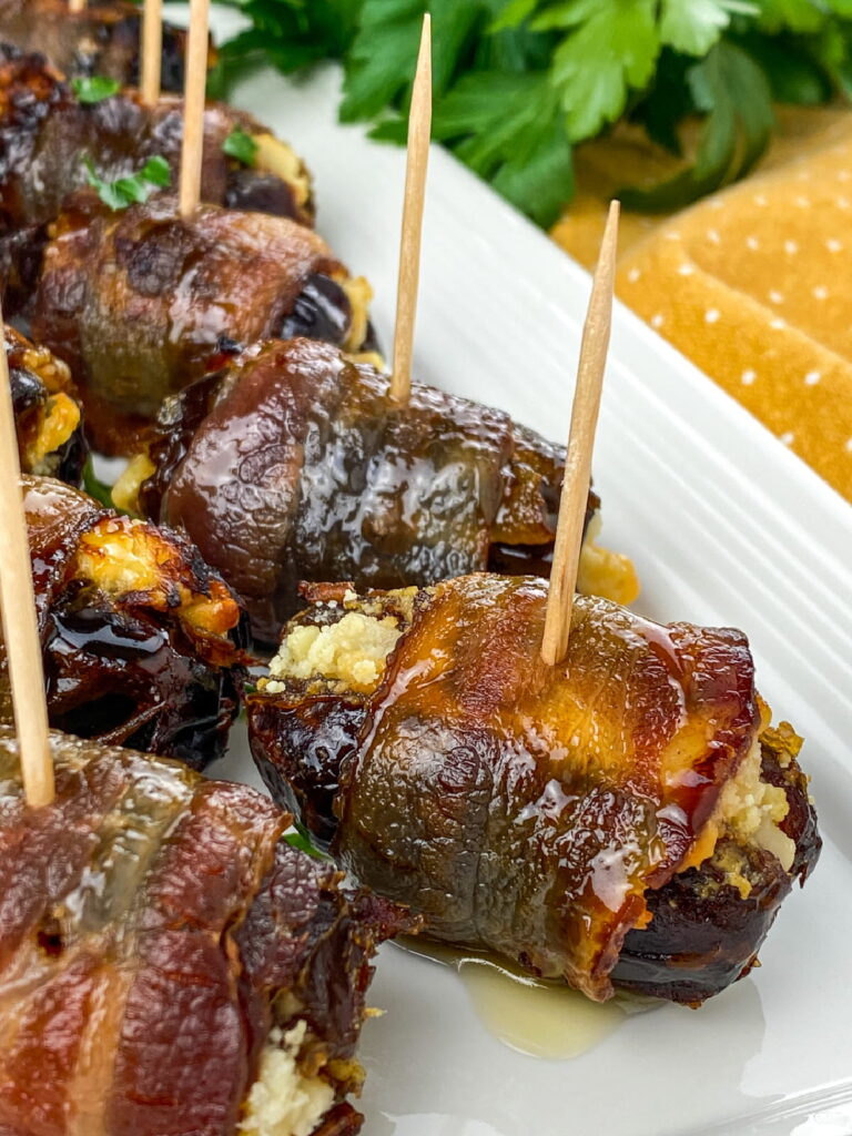 Bacon-wrapped stuffed dates secured with toothpicks are arranged on a white plate, garnished with herbs.