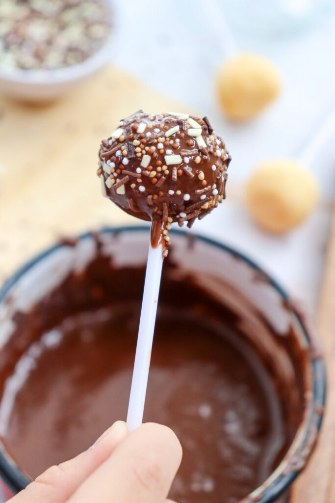 A hand holding a cake pop covered in chocolate and sprinkles above a bowl of melted chocolate.