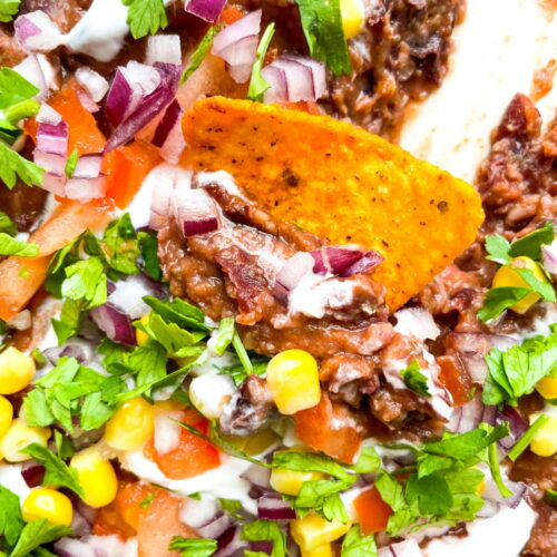Close-up of a tortilla chip in a dip with beans, corn, tomato, red onion, cilantro, and sour cream.